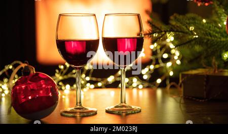 Christmas celebration. Two red wine glasses and decoration on table, burning fireplace background. Xmas holiday Stock Photo