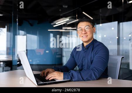 Successful asian businessman smiling and looking at camera, programmer porter inside office using laptop writing code for software security. Stock Photo