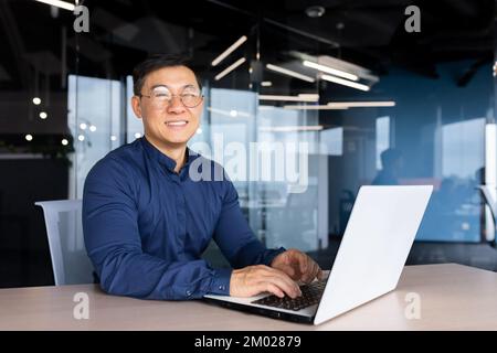 Successful asian businessman smiling and looking at camera, programmer porter inside office using laptop writing code for software security. Stock Photo