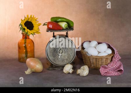 A fine art still life photograph of a dozen eggs in a basket, vegetables on an old scale, and a sunflower in a vintage bottle. Stock Photo