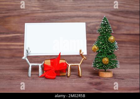 Tiny deer figurines holding a blank wish card by a toy Christmas tree and wrapped gift box against a dark wooden background. Creative holidays season Stock Photo