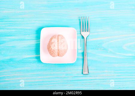 Miniature toy human brain model on a tiny white plate and small silver fork isolated on blue background. Mind diet for brain health. Stock Photo