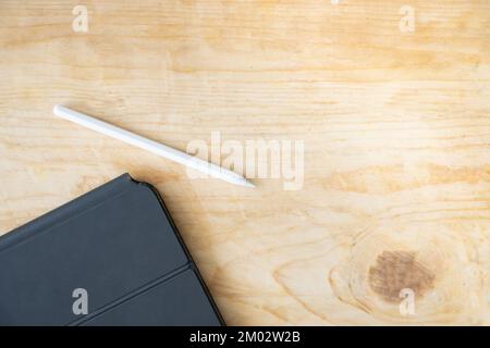 White digital stylus pen beside a tablet in a dark protective case on a light wooden table. Top view with copy space for creative design. Stock Photo