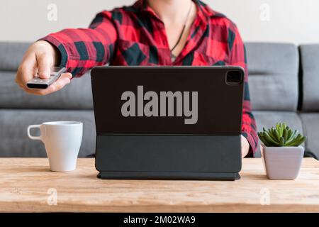 Person pointing a remote control toward the camera with a tablet, white mug and small plant on a wooden table at home. Stock Photo
