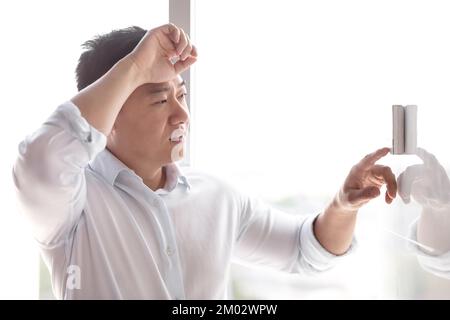 Close-up photo. A young Asian man tired from the heat in a white shirt turns on the control panel of the air conditioner on the wall with his finger. Holds his head, wipes sweat. Stock Photo