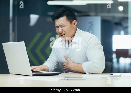 Heart attack at the workplace. A young Asian man is sitting in the office at the table, holding his chest, grimacing, feeling pain, having a stroke. Stock Photo