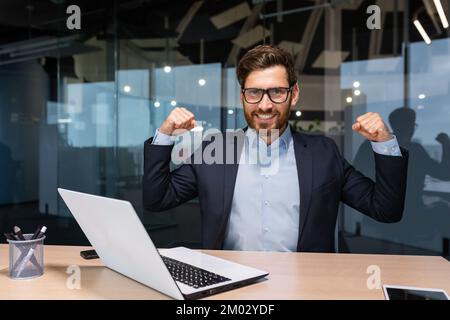 Portrait of mature senior businessman in office, man looking at camera and holding hands up super power and superhero gesture, successful investor using laptop inside building. Stock Photo