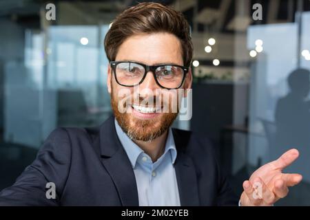 Businessman looks into smartphone camera and talks on video call, man uses phone for remote communication with friends and colleagues, investor boss in glasses works inside office. Stock Photo
