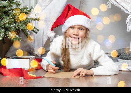 Christmas miracle wish list. Smiling girl in Santa Claus hat and a white sweater writing letter dreams for gifts to Santa Claus. Child lies at home. Stock Photo