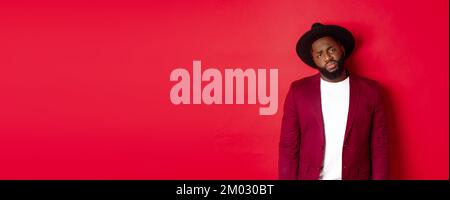 Sad and unamused Black man looking with pity at camera, frowning disappointed, standing against red background Stock Photo