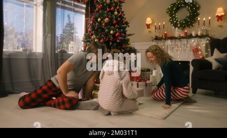 Young girl opens gifts from parents and rejoicing on Christmas morning under big decorated Christmas tree. Warm atmosphere at home on Christmas or New Year. Happy childhood. Winter holidays. Stock Photo