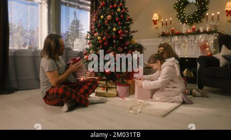 Young girl opens gifts from parents and rejoicing on Christmas morning under big decorated Christmas tree. Warm atmosphere at home on Christmas or New Year. Happy childhood. Winter holidays. Stock Photo