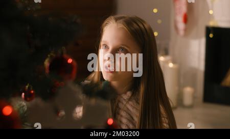 Young girl in mild pajama sits on the floor, decorates Christmas tree with balls and toys, smiles and spends leisure time at home. Child prepares room before winter holidays and New Year. Slow motion. Stock Photo