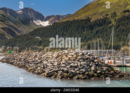 Seward, Alaska, USA - July 22, 2011: Gray rocky wave breaker as protection of the harbor with masts peeping over it. Gray mountain with green forested Stock Photo