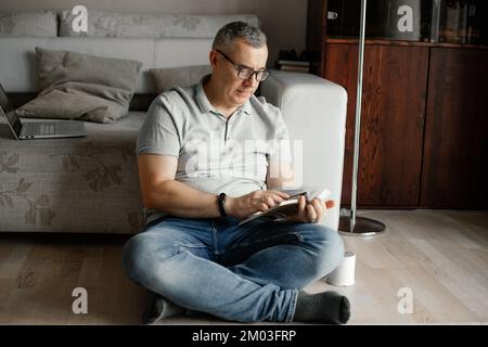 Portrait of concentrated middle-aged man sitting with crossed legs near sofa on floor, holding reading book at home. Stock Photo