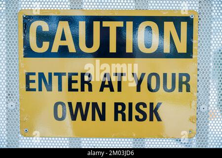 Caution Enter at Your Own Risk on a yellow sign- Miami, Florida. Close-up of a warning sign posted on a white screen fence at the background. Stock Photo