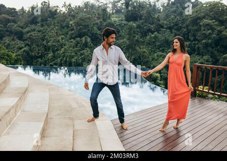 Portrait of young couple in love walking by pool at the resort. Young man and woman holding hand walking near swimming pool of holiday resort. Stock Photo