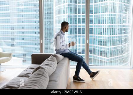 Thoughtful dreamy millennial Black man using smartphone in office lobby Stock Photo
