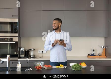 Joyful attractive young Black chef man holding smartphone, looking away Stock Photo