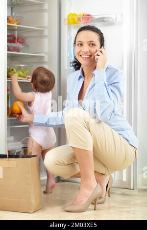 You have to have eyes in the back of your head. a mother talking on the phone while putting groceries away with her baby rummaging through the fridge. Stock Photo