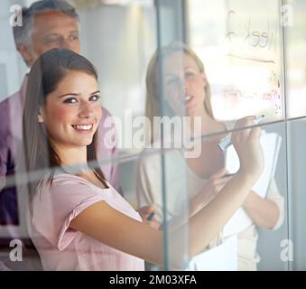 Our strategy works. A young businesswoman writing down plans on a glass pane while her associates look on. Stock Photo