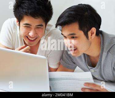 Doing a little house-hunting online. a young gay couple relaxing with their laptop in bed. Stock Photo