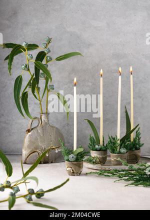 Beautiful winter floristic arrangement with white narrow candles, eucalyptus leaves and fruits in four ceramic mugs. Handmade Christmas home decor Stock Photo