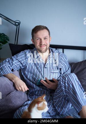 Attractive caucasian bearded man using tablet computer sitting in bed in sleepwear making purchases online, working or surfing while tabby cat is sitting with him. High quality vertical image Stock Photo