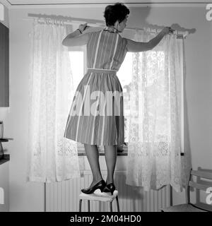 1960s home decorating. A woman at home putting up the curtains in the room window. All by herself she balances on a chair and a stool to manage it. Sweden 1966 Stock Photo