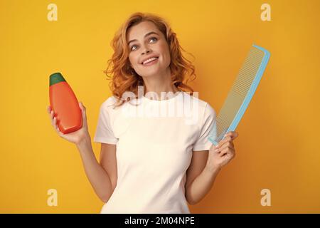 Surprised amazed girl combing hair. Redhead woman with a comb, isolated on yellow background. Woman hold bottle shampoo and conditioner. Stock Photo