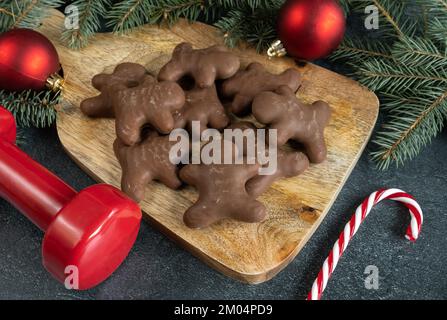 Chocolate gingerbread cookies, gym dumbbell, candy cane, Christmas baubles decorations on tree branches. Winter fitness diet, workout dieting concept. Stock Photo