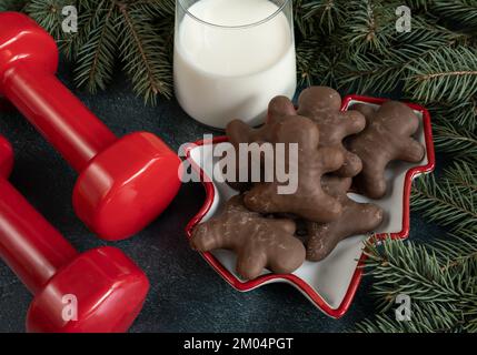 Dumbbells, chocolate gingerbread cookies, glass of milk for Santa Claus, Christmas tree branches. Fitness winter diet, gym workout, dieting concept. Stock Photo