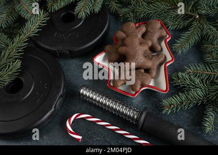 Gym dumbbell barbell weight plates, chocolate gingerbread Christmas cookies, candy cane, tree branches. Winter fitness diet, workout dieting concept. Stock Photo