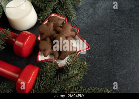 Dumbbells, Christmas cookies, glass of milk for Santa Claus, tree branches. Winter diet composition. Gym workout, dieting concept with copy space. Stock Photo