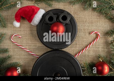 Dumbbells barbell weight plates shaped as snowman in red Santa Claus Christmas hat. Fitness holiday season winter composition. Gym workout concept. Stock Photo