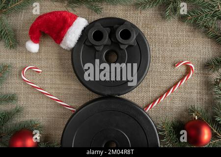 Dumbbells barbell weight plates shaped as snowman in red Santa Claus Christmas hat. Fitness holiday season winter composition. Gym workout concept. Stock Photo