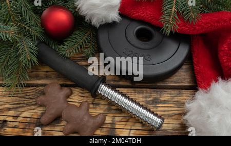 Dumbbell barbell weight plate, chocolate gingerbread Christmas cookies, red Santa Claus hat. Winter fitness diet, gym workout, dieting concept. Stock Photo