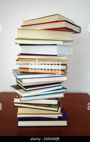 A pile of books of different shapes and thicknesses on a wooden shelf and a white wall in the background Stock Photo