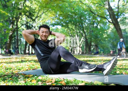 Portrait of a young handsome Asian man doing sports exercises, yoga, sports in the park on a mat. Leads a healthy lifestyle. He looks at the camera, smiles. Stock Photo