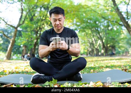 Young handsome male Asian sportsman sits in the park on a mat in black sportswear and uses a mobile phone. Resting after active training. Stock Photo