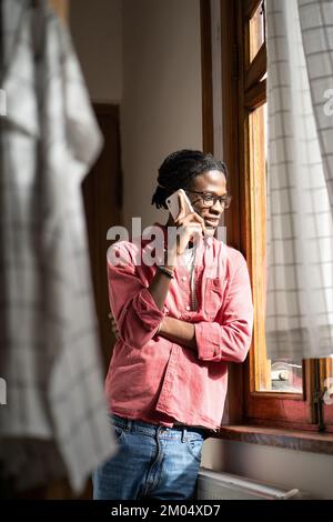 Smiling millennial African guy talking on phone with girlfriend, standing by window at home Stock Photo