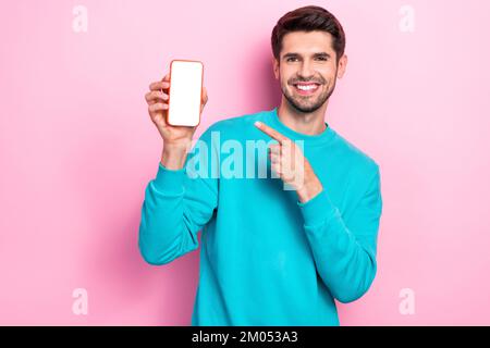Photo of young toothy beaming smile guy student wear blue sweater hold phone finger direct empty space touchscreen isolated on pink color background Stock Photo