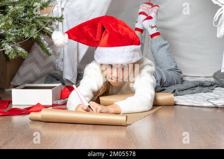 Christmas miracle wish list. Christmas helper child writing letter to Santa Claus in red hat. smiling girl making wish list or letter to santa at home Stock Photo