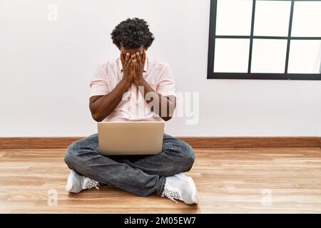 African man with curly hair using laptop sitting on the floor with sad expression covering face with hands while crying. depression concept. Stock Photo