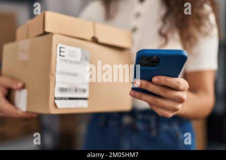 Young beautiful hispanic woman ecommerce business worker using smartphone holding package at office Stock Photo