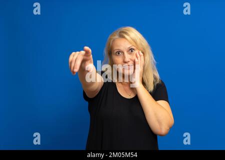 Surprised attractive cute blonde straight-haired caucasian woman in casual black t-shirt pointing finger at the far road. Isolated on blue background. Stock Photo