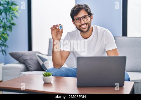 Handsome latin man holding virtual currency bitcoin using laptop looking positive and happy standing and smiling with a confident smile showing teeth Stock Photo