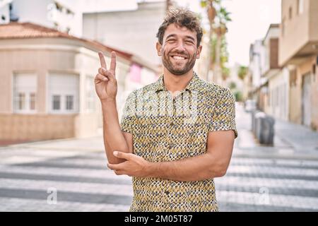 Young hispanic man standing at the street smiling with happy face winking at the camera doing victory sign. number two. Stock Photo