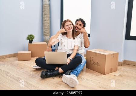 Young couple using laptop at new home smiling doing phone gesture with hand and fingers like talking on the telephone. communicating concepts. Stock Photo