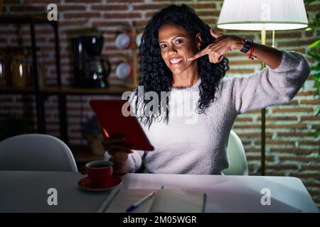 Middle age hispanic woman using touchpad sitting on the table at night smiling cheerful showing and pointing with fingers teeth and mouth. dental heal Stock Photo